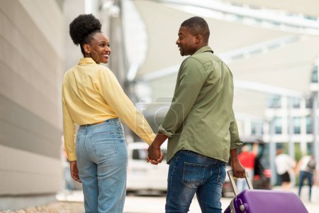 Photo for Cheerful Black Spouses Traveling On Vacation Walking With Travel Suitcase At Modern Airport Outside. Married Couple Holding Hands Entering Terminal. Transportation Concept - Royalty Free Image