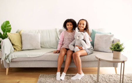 Foto de Dos hermanas negras abrazando posando con un juguete de oso esponjoso sentado en el sofá en la sala de estar moderna en interiores. Una foto de dos amiguitas en casa. Concepto de Infancia y Amistad - Imagen libre de derechos