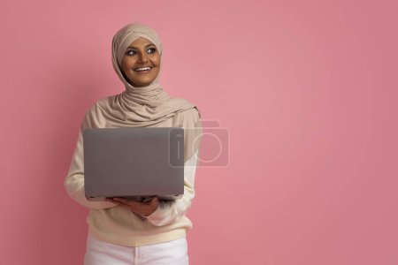 Photo for Online Education. Portrait of smiling muslim female in hijab holding laptop and looking aside, islamic woman enjoying distance learning, posing with computer in hands on pink studio backgrond - Royalty Free Image