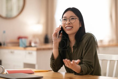 Téléchargez les photos : Joyeux heureux jolie jeune femme asiatique portant des lunettes entrepreneur assis à la tablette de cuisine, avoir un appel téléphonique avec le client, en regardant l'espace de copie et des gestes, travailler à la maison - en image libre de droit