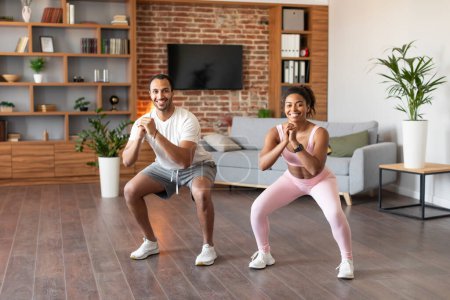 Photo for Satisfied young black woman and male athletes in sportswear squat, enjoy workout together at spare time in living room interior. Body care, slimming and fitness for muscles at home during covid-19 - Royalty Free Image