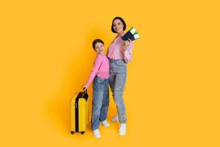 Photo for Family Travel. Happy Young Mother And Daughter With Tickets And Suitcase Posing Over Yellow Studio Background, Cheerful Mom And Female Child Ready For Vacation Trip, Full Length, Copy Space - Royalty Free Image