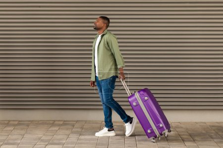 Téléchargez les photos : Homme afro-américain souriant marchant avec une valise de voyage regardant à l'extérieur près du mur de l'aéroport. Un type qui voyage en vacances. Offre tour et transport. Longueur totale - en image libre de droit