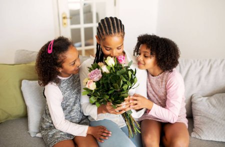Photo for Mothers Day Celebration. Black Daughters Giving Flowers To Mom Greeting Her Celebrating Holiday Sitting On Sofa At Home. Young Mommy Holding Bouquet Posing With Kids In Modern Living Room - Royalty Free Image