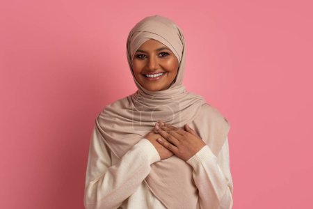 Téléchargez les photos : Concept de gratitude. Portrait de femme musulmane reconnaissante en hijab en gardant les mains sur la poitrine et en regardant la caméra, souriant dame arabe exprimant son appréciation, debout sur fond de studio rose - en image libre de droit