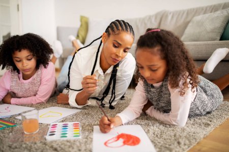 Téléchargez les photos : Mère afro-américaine tressée et deux filles dessinant un cœur à colorier, faisant peindre ensemble allongé sur le sol dans le salon moderne à l'intérieur le week-end. Loisirs et loisirs en famille. Focus sélectif - en image libre de droit