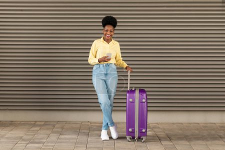 Téléchargez les photos : Grande demande de voyage. Happy Black Woman Traveler Utilisation téléphone réservation billets d'avion en ligne debout avec valise à l'extérieur, sourire à la caméra. Prise de vue complète. Offre vacances Concept - en image libre de droit
