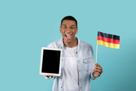 Téléchargez les photos : Heureux choqué homme noir millénaire excité dans occasionnel montrant drapeau de l'Allemagne et tablette avec écran vide isolé sur fond bleu studio. App recommandation pour apprendre l'allemand, étude d'échange - en image libre de droit