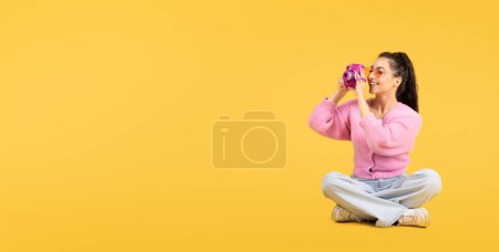 Téléchargez les photos : Joyeux jeune femme brune caucasienne en lunettes de soleil avec appareil photo faire des photos, s'assoit sur le sol, regarde l'espace libre isolé sur fond de studio jaune, panorama. Annonce et offre de voyage et vacances - en image libre de droit