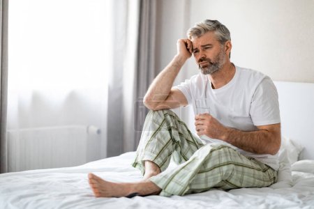 Téléchargez les photos : Malheureux homme d'âge moyen en pyjama souffrant de maux de tête matinaux après avoir dormi dans la chambre, assis sur le lit après s'être réveillé, toucher la tête, tenant un verre d'eau, regardant l'espace de copie - en image libre de droit