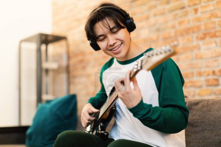 Photo for Cheerful Asian Student Guy Playing Electric Guitar Wearing Earphones Sitting On Sofa In Modern Living Room At Home. Boy Learning New Chords On Weekend. Hobby And Musical Gadgets. Selective Focus - Royalty Free Image