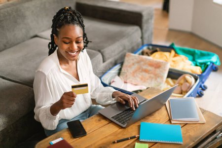 Foto de Mujer afroamericana feliz utilizando la tarjeta de crédito y el ordenador portátil, reservar en el extranjero viaje, navegar por el sitio web de la agencia de viajes en línea o hacer la reserva de hotel en la web desde casa - Imagen libre de derechos