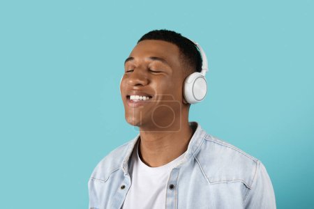 Photo for Glad millennial black male in wireless headphones with closed eyes listen music, enjoy audio app and spare time isolated on blue studio background, close up. Ad and offer, great mood, relax - Royalty Free Image