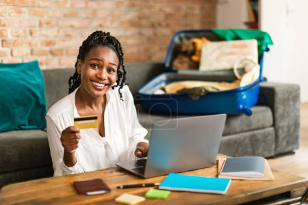 Foto de Emocionado afroamericano señora reservar vuelo en línea con ordenador portátil y tarjeta de crédito, sentado con maleta desempacada en casa. Mujer comprando entradas baratas en la web - Imagen libre de derechos