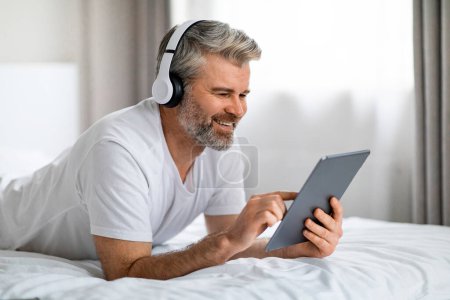 Téléchargez les photos : Charmant homme d'âge moyen barbu aux cheveux gris positif en vêtements de maison couché sur le lit à la maison, utilisant des écouteurs sans fil modernes et une tablette numérique, écran tactile, choisissant le contenu vidéo en ligne, espace libre - en image libre de droit