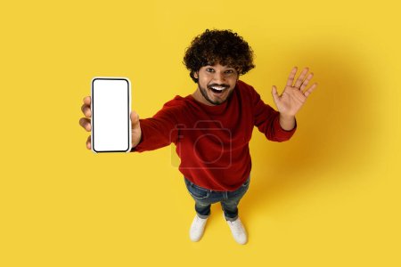Téléchargez les photos : Vue grand angle de joyeux positif millénial beau mec indien cool montrant smartphone avec écran blanc vierge, maquette pour la publicité grande offre, fond de studio jaune - en image libre de droit