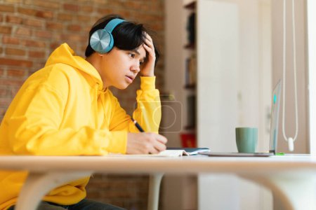 Photo for Hardworking Korean Teenager Boy Looking At Laptop Learning Online Wearing Wireless Earphones Sitting At Desk At Home. Tired Guy Doing Homework Studying Remotely. Side View Shot - Royalty Free Image
