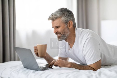 Photo for Relaxed cheerful happy handsome mature grey-haired man lying on bed at home, using modern pc laptop, typing on computer keyboard and smiling, drinking coffee, scrolling on Internet, copy space - Royalty Free Image