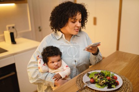 Photo for Mom blog. Hispanic curly young lady plus size mother sitting at kitchen table, holding baby toddle on her lap while have lunch, using smartphone, mommy blogger taking photo of her fresh healthy salad - Royalty Free Image