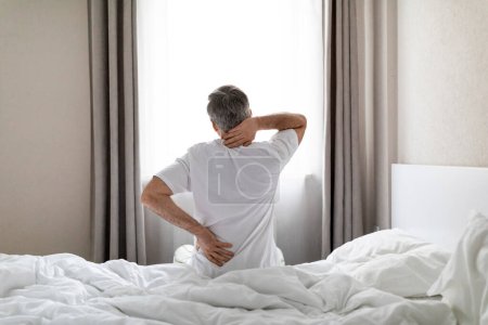 Photo for Unrecognizable mature grey-haired man wearing white pajamas sittting on bed at home in the morning, touching lower back and neck, suffering from muscle strain after waking up, copy space - Royalty Free Image
