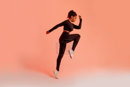 Photo for Athletic black lady jumping in mid air, training over peach neon studio background, full length, side view, copy space. Workout and sport motivation concept - Royalty Free Image