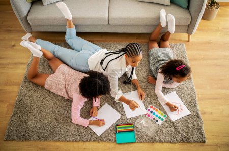 Photo for Above View Of African American Family Of Three Drawing And Having Fun Lying On Floor At Home. Mother And Two Daughters Painting Spending Time Together On Weekend. Hobby And Talent - Royalty Free Image