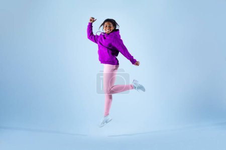 Téléchargez les photos : Femme noire joyeuse en tenue de sport s'amusant à sauter haut sur fond bleu, plan studio, pleine longueur. Athlète énergique en vêtements de sport et baskets sautant et souriant - en image libre de droit
