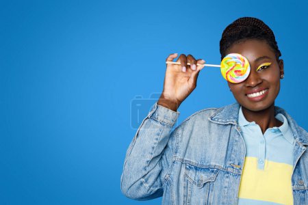 Téléchargez les photos : Joyeux ludique belle jeune femme noire en tenue décontractée avec des fards à paupières lumineux couvrant un oeil avec des bonbons sucette colorée et souriant à la caméra, posant sur fond bleu, espace de copie - en image libre de droit