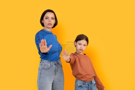 Foto de Retrato de la madre y su hijita mostrando el gesto de stop en la cámara, la joven confiada y su hijo preadolescente de pie aislado sobre el fondo amarillo en el estudio, espacio de copia - Imagen libre de derechos