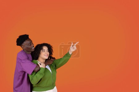 Téléchargez les photos : Câlins affectueux gai jeune couple mixte dans occasionnel afro-américain homme et hispanique chubby femme pointant vers l'espace de copie pour la publicité sur fond de studio rouge, montrant beaucoup - en image libre de droit