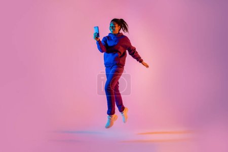 Téléchargez les photos : Plan complet de la longueur du corps de jeune femme noire en sautant de vêtements de sport et en utilisant un téléphone portable sur fond rose néon couleur claire, espace de copie gratuit - en image libre de droit