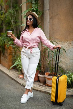 Photo for Cheerful young black lady in casual, sunglasses with suitcase enjoy trip in new city with cup of coffee, outside, vertical, free space. Vacation and travel at summer, coffee break - Royalty Free Image