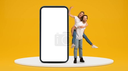 Photo for Glad young european man in white t-shirt hold lady on back, female shows peace sign with hand with huge phone with blank screen, look at camera isolated on yellow background. Couple have fun, app - Royalty Free Image