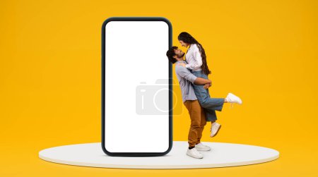 Téléchargez les photos : Heureux millénial indien gars tenir petite amie, amusez-vous près de téléphone énorme avec écran blanc sur fond de studio jaune, profil, espace libre. Romantique, amour, app et réseaux sociaux - en image libre de droit