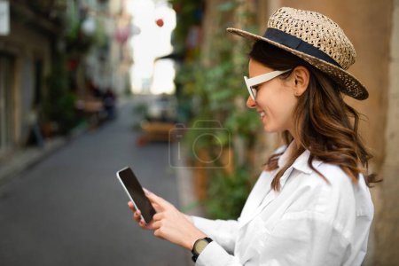 Téléchargez les photos : Joyeux jeune femme caucasienne en casual et chapeau fait photo sur smartphone avec écran vide en ville, à l'extérieur. Appareil, application pour les vacances et blog de voyage en été, appel vidéo et réseaux sociaux - en image libre de droit