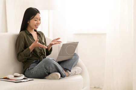 Téléchargez les photos : Joyeux positif jolie jeune femme chinoise dans occasionnel assis sur le canapé avec les jambes vers le haut, avoir une réunion en ligne avec des amis, en utilisant un ordinateur portable, en regardant l'écran d'ordinateur, souriant et gestuel, copier l'espace - en image libre de droit
