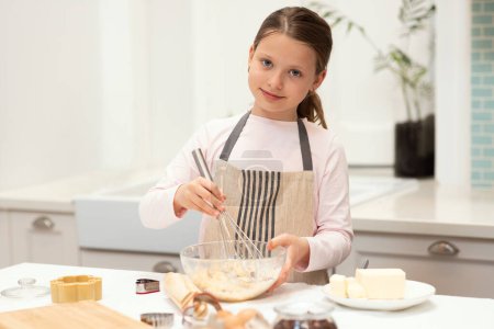 Téléchargez les photos : Joyeux caucasien petite fille dans tablier faire pâte à biscuits avec fouet à l'intérieur de la cuisine, copier l'espace. Cuisson de bonbons, nourriture maison, culinaire à temps libre, préparer à manger à la maison - en image libre de droit