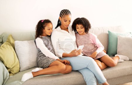 Foto de Feliz madre afroamericana y dos hijas usando el teléfono juntas viendo dibujos animados en línea sentadas en el sofá en casa. Padres e hijos navegando por Internet a través de Smartphone. Familia Ocio Y Gadgets - Imagen libre de derechos