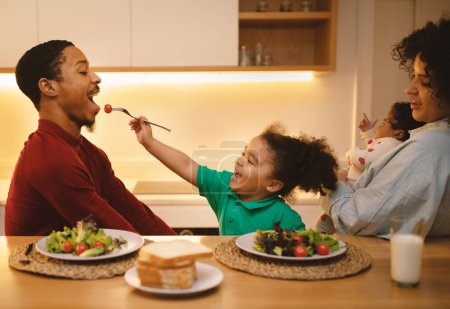 Téléchargez les photos : Belle famille interracial heureuse et aimante déjeuner à la cuisine moderne à la maison. Mignon bébé fille nourrir son père homme afro-américain, hispanique bouclée femme tenant bébé, profiter du temps ensemble - en image libre de droit