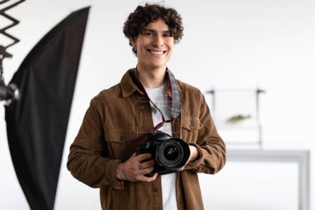 Téléchargez les photos : Joyeux jeune homme photographe tenant un appareil photo professionnel, travaillant dans un studio de photographie moderne, regardant et souriant à la caméra, espace libre - en image libre de droit