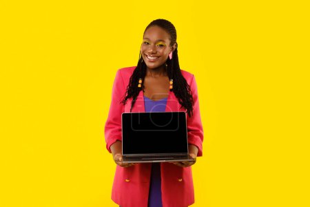 Téléchargez les photos : Femme noire joyeuse montrant ordinateur portable avec la publicité d'écran vide et recommandant le site Web en ligne debout sur fond jaune dans Studio, souriant à la caméra. Maquette - en image libre de droit