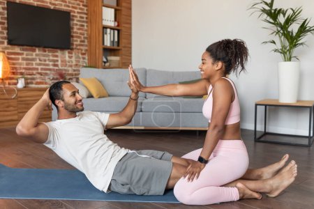 Photo for Happy young black lady helping guy in sportswear doing abs exercises on mat on floor together, holds hands in living room interior. Body care with support and help, weight loss and fitness at home - Royalty Free Image