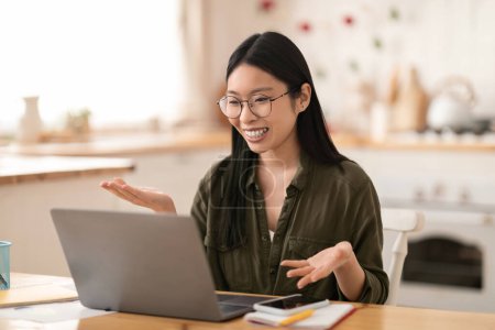 Téléchargez les photos : Amical positive jolie jeune femme asiatique dans occasionnel assis à la table de cuisine, avoir une réunion en ligne avec des partenaires d'affaires, à l'aide d'un ordinateur portable, en regardant l'écran d'ordinateur, souriant et gestuel, copier l'espace - en image libre de droit