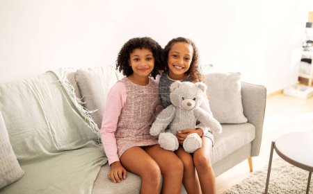 Téléchargez les photos : Deux sœurs afro-américaines mignonnes étreignant tenant peluche jouet ours assis sur le canapé posant ensemble portant des robes décontractées à la maison, souriant à la caméra. Frères et sœurs Concept d'amitié - en image libre de droit