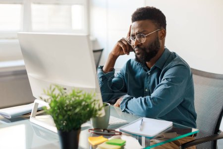 Téléchargez les photos : Homme noir réfléchi PDG travaillant sur ordinateur au bureau, assis sur le lieu de travail et pensant à un projet d'entreprise à l'intérieur. Carrière d'entreprise et nouveau concept de start-up - en image libre de droit