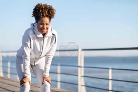 Téléchargez les photos : Femme noire sportive prenant une pause pendant l'entraînement à l'extérieur, penchant les mains sur les genoux, femme afro-américaine athlétique en vêtements de sport se détendre après l'entraînement extérieur, debout sur une jetée en bois près de la mer - en image libre de droit