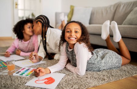 Téléchargez les photos : Heureuse petite fille noire dessinant avec mère et sœur ensemble, collant et s'amusant allongé sur le sol dans le salon à la maison, souriant à la caméra. Week-end familial Loisirs Concept. Profondeur faible - en image libre de droit