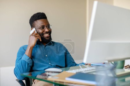 Photo for Happy black businessman having phone conversation with client, sitting in front of computer at office and talking on cellphone. Modern business concept - Royalty Free Image