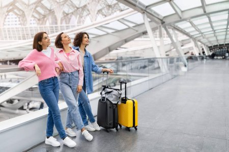 Téléchargez les photos : Trois belles amies debout à l'aéroport et regardant loin, Groupe de jeunes femmes heureuses avec des valises à bagages en attente d'un vol au Terminal Hall, Profitant de voyager ensemble, Copier l'espace - en image libre de droit