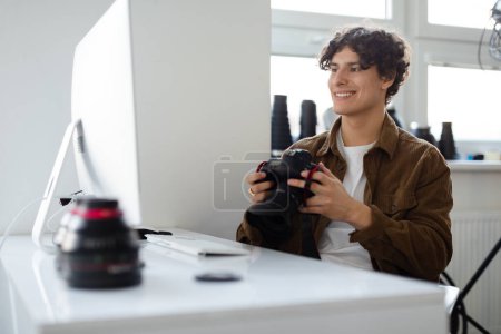 Téléchargez les photos : Joyeux photographe professionnel tenant la caméra et regardant le moniteur d'ordinateur, assis sur le lieu de travail en studio. Artiste professionnel transférant des images à l'ordinateur pour retoucher - en image libre de droit